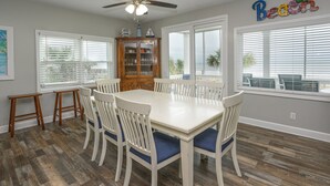 Un coin repas avec une table et des chaises blanches, un buffet en bois et de grandes fenêtres équipées de stores.