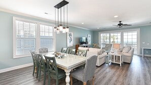 A spacious living room with a dining area, large windows, and a ceiling fan.
