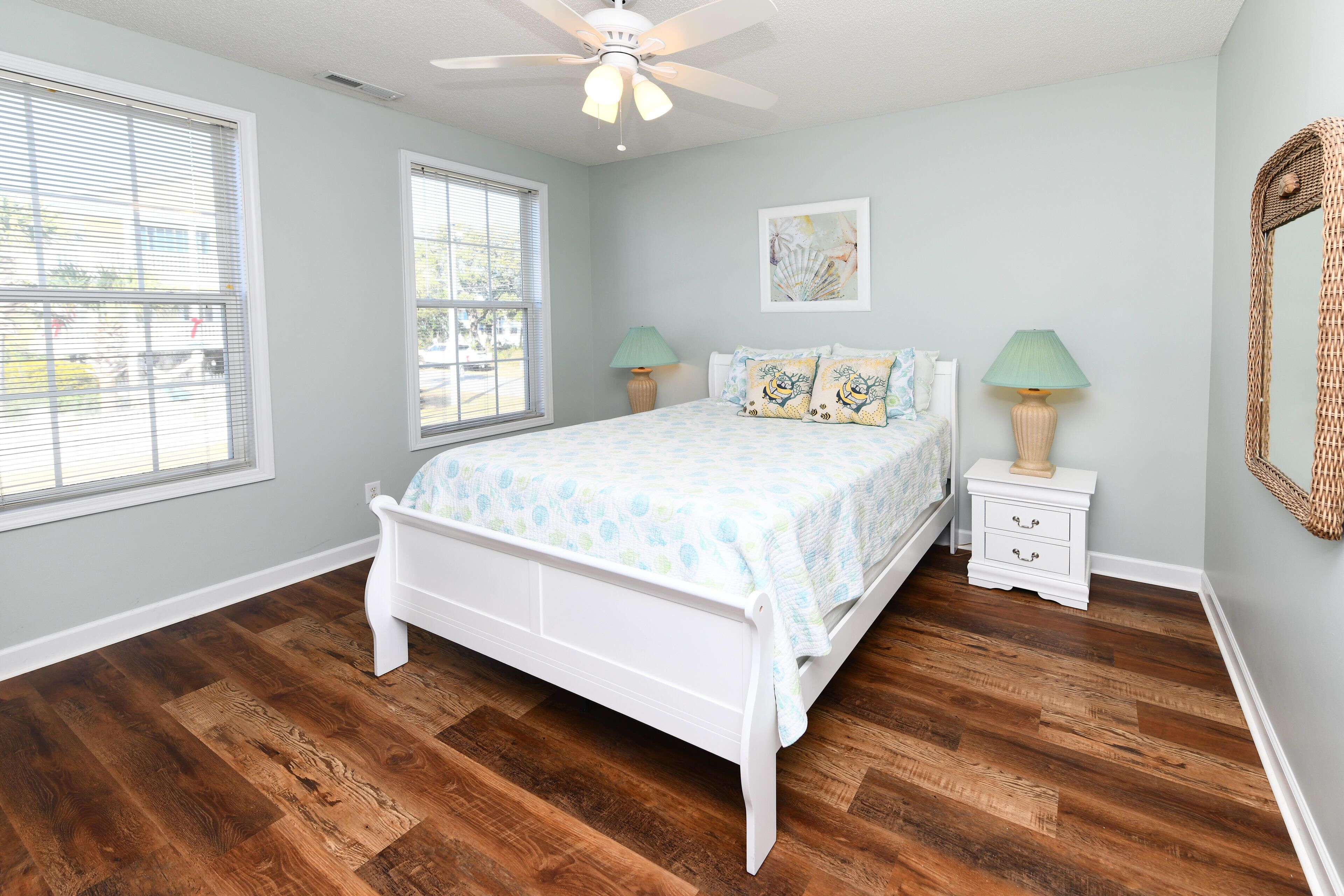 A bedroom with a white bed, two bedside tables with lamps, a ceiling fan, and large windows.