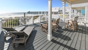 A wooden deck with lounge chairs and a table, overlooking a beach and pier.