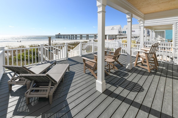 A wooden deck with lounge chairs and a table, overlooking a beach and pier.