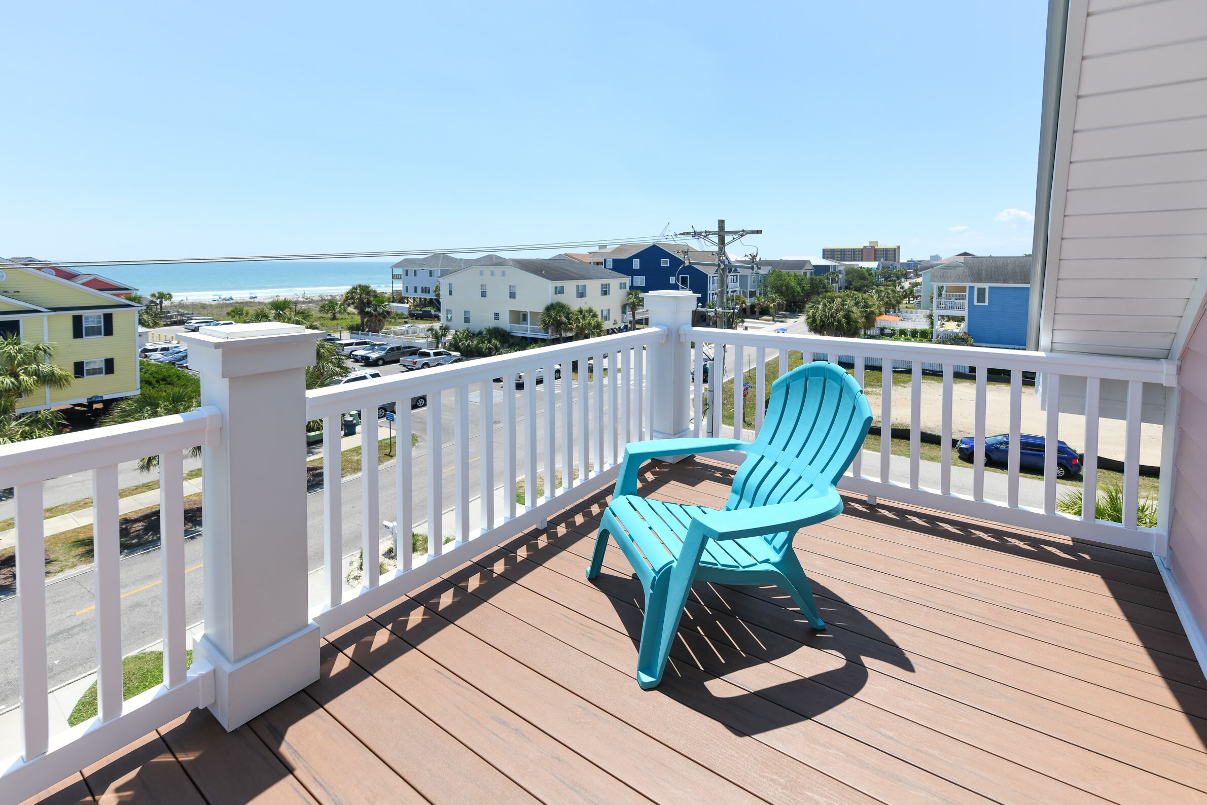 Um deck de madeira com duas cadeiras Adirondack turquesa, corrimãos brancos e vista para a praia e edifícios.