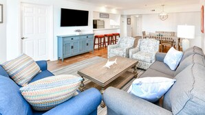 Un salon avec un canapé bleu, des coussins à motifs, une table basse en bois et un canapé gris. Un téléviseur est fixé au mur, et on aperçoit un coin cuisine au fond, ainsi qu’une table à manger avec des chaises.