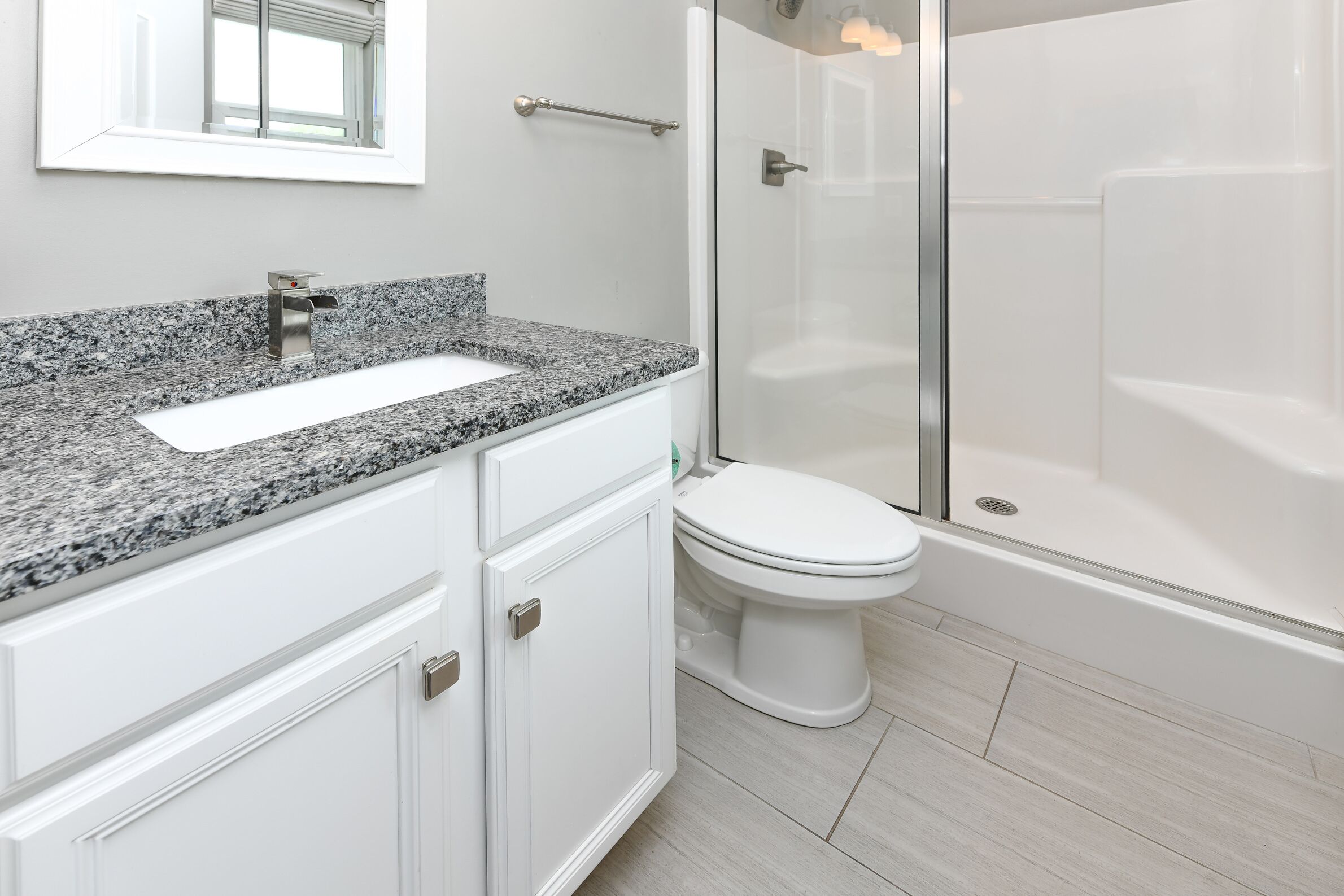 A bathroom with a granite countertop sink, white cabinets, a toilet, and a glass-enclosed shower.