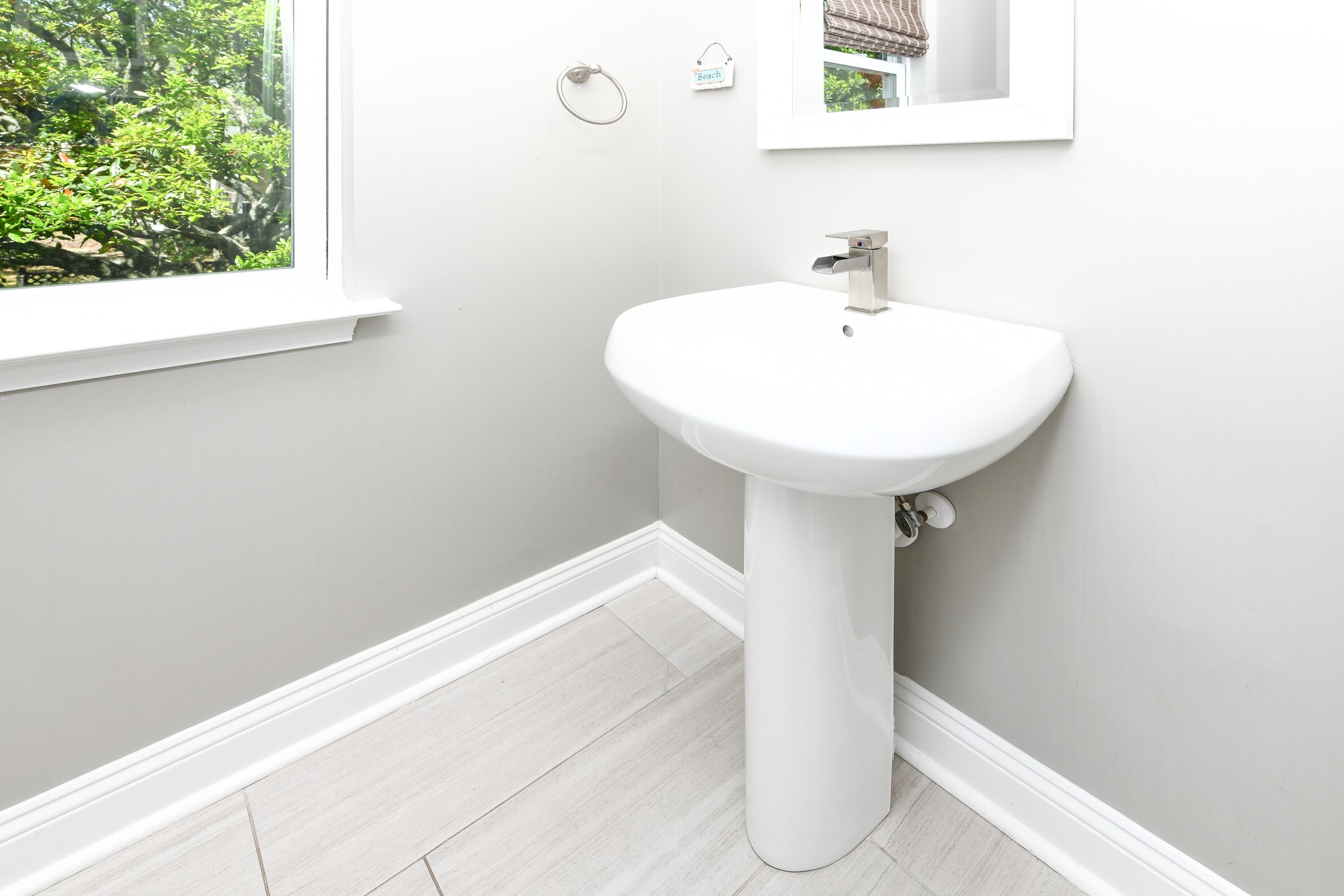 Une salle de bain avec un lavabo sur colonne blanc, une fenêtre donnant sur un espace verdoyant et un petit panneau accroché au mur.