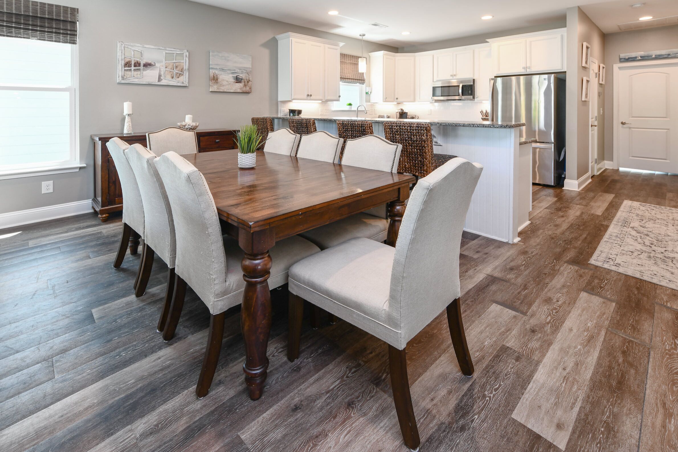 A dining area with a wooden table and upholstered chairs, a kitchen with white cabinets and stainless steel appliances, and a herringbone-patterned hardwood floor.