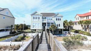 A two-story house with a wooden deck leading to the front door, surrounded by a white fence and palm trees. - Oceanfront 7 BR Beach House with a Private Pool! Royal Palm (Garden City)