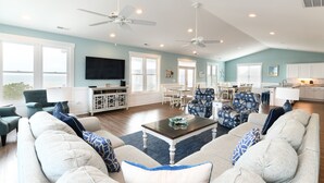 A spacious living room with a large sofa, a coffee table, and a dining area with chairs. There are ceiling fans, a TV mounted on the wall, and a view of the ocean through large windows. - Ocean View 7 BR Beach House with a Private Pool! Seaside Retreat  (Surfside Beach)