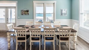 A dining room with a large table and six chairs, a view of the ocean through large windows, and a door leading outside. - Ocean View 7 BR Beach House with a Private Pool! Seaside Retreat  (Surfside Beach)