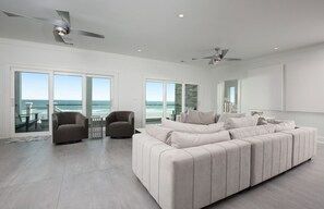 Un salón amplio con un sofá seccional beige grande, dos sillones y una mesa de vidrio. La habitación tiene puertas correderas de vidrio que dan a un balcón con vistas al océano.