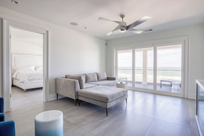 Uma sala de estar espaçosa com um sofá grande, uma mesa de centro e um ventilador de teto. Há uma janela ampla com vista para a praia.