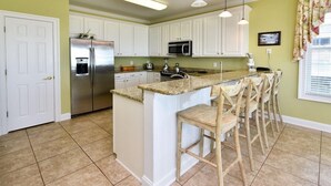 Una cocina con gabinetes blancos, un refrigerador de acero inoxidable, un microondas y una isla de encimera de granito con taburetes de bar.