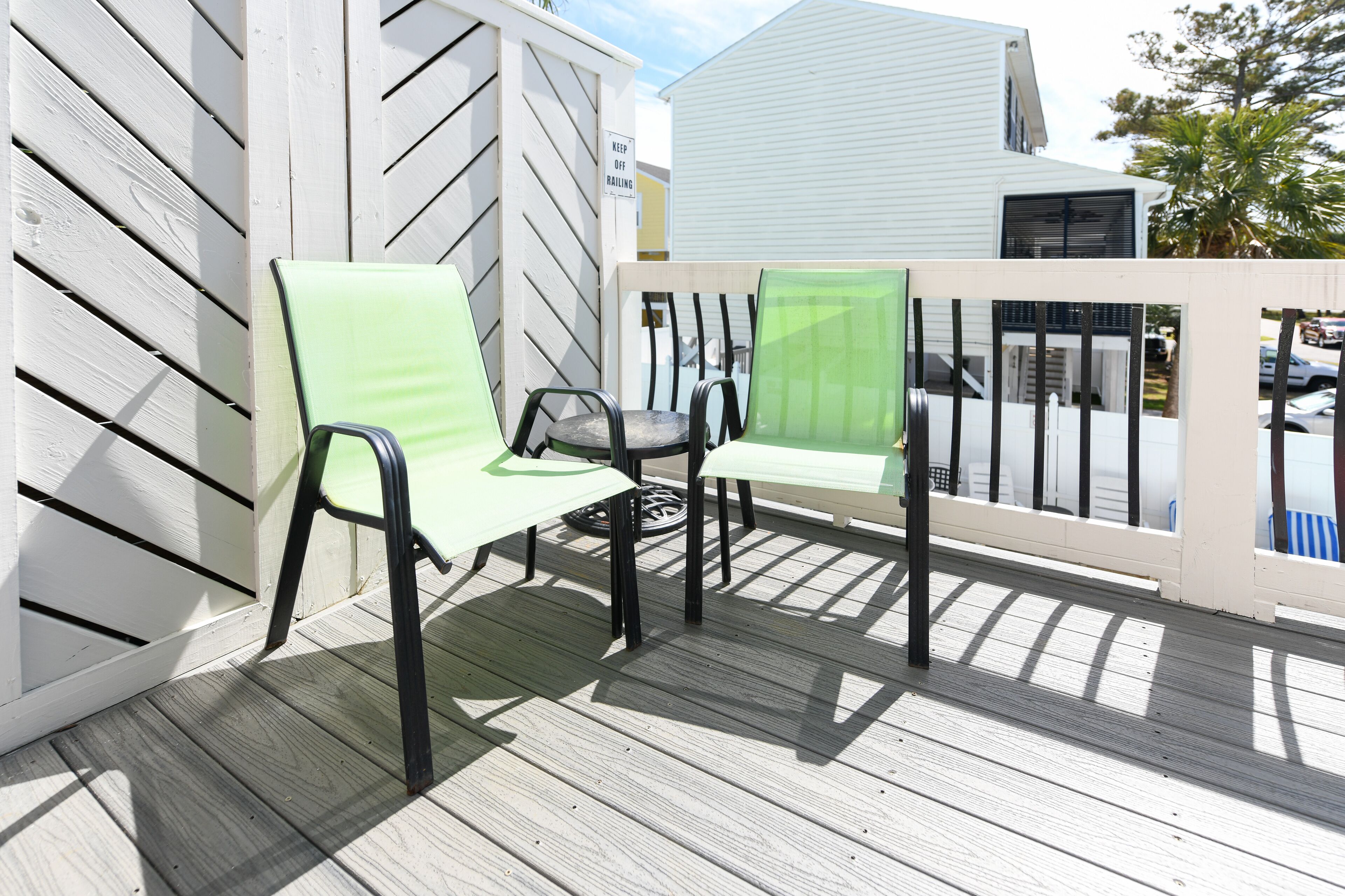 Two outdoor chairs on a wooden deck with a white fence and a sign that reads 'KEEP OFF RAILING'.