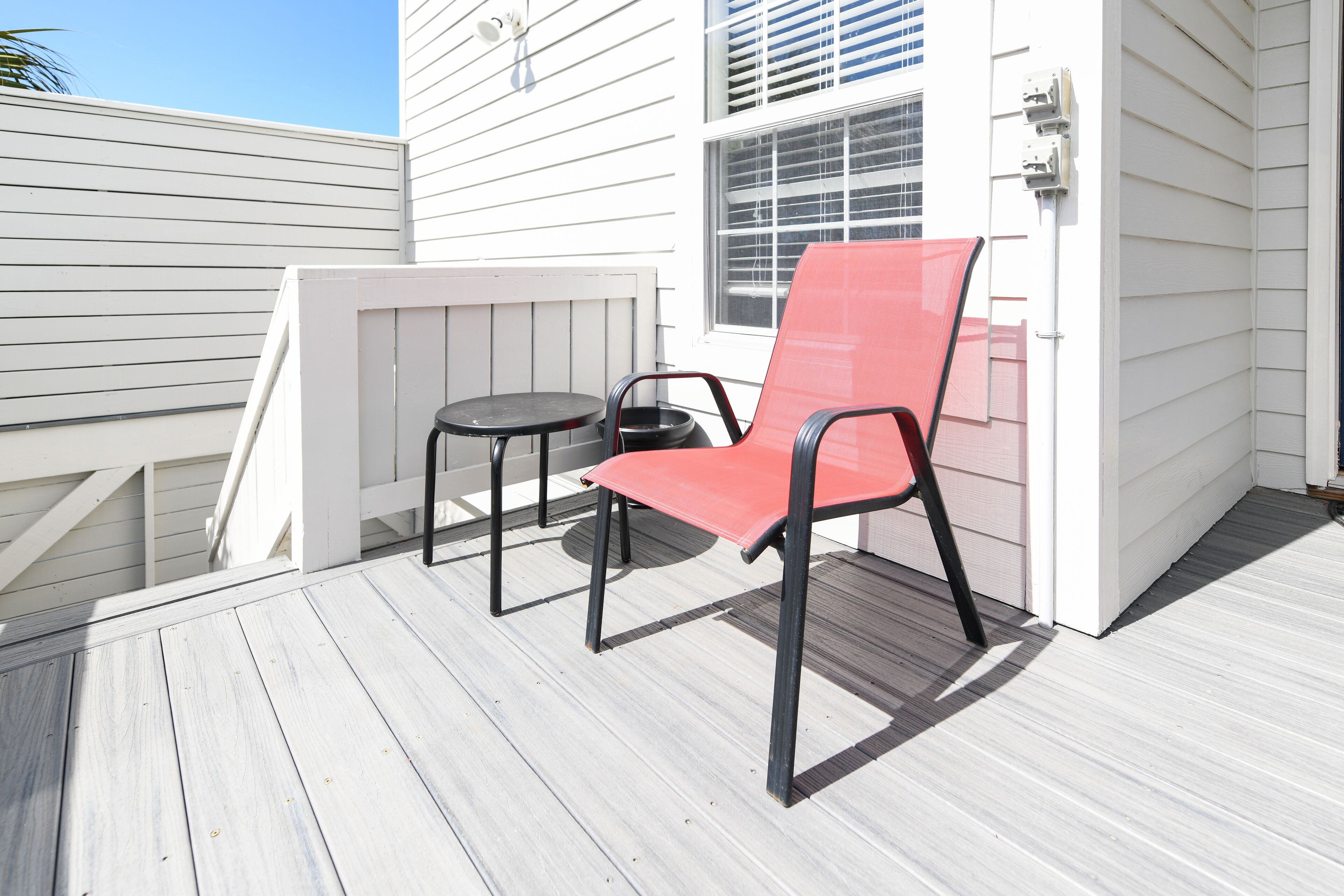 Une terrasse avec une chaise rouge et une petite table ronde, un revêtement extérieur blanc et une fenêtre avec des stores.
