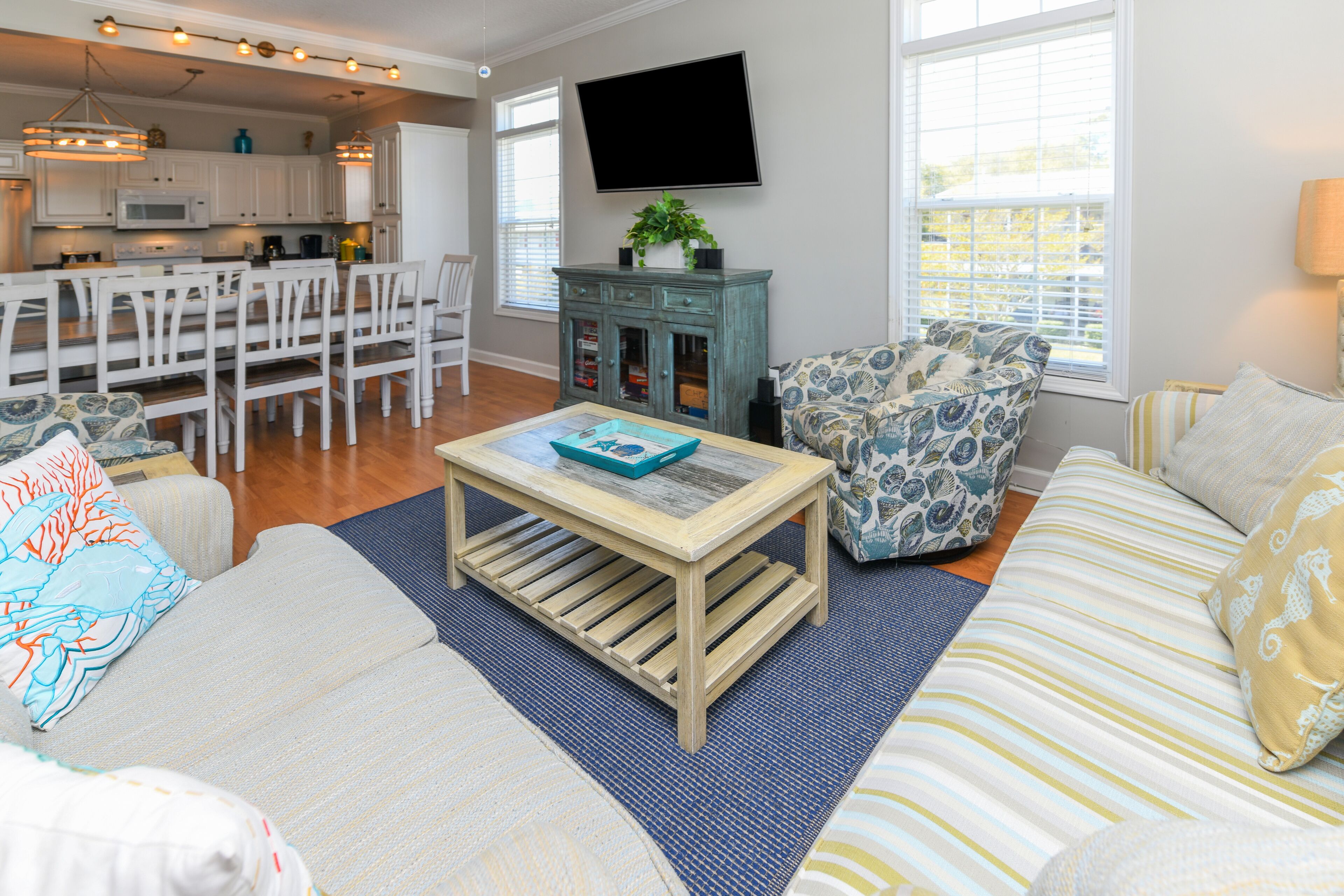 A modern living room with a sofa, coffee table, and patterned armchairs. A flat-screen TV is mounted on the wall, and a kitchen is visible in the background.