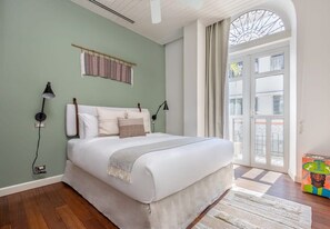 A bedroom with a large bed, a window with curtains, a wall-mounted shelf, and a bedside lamp.
