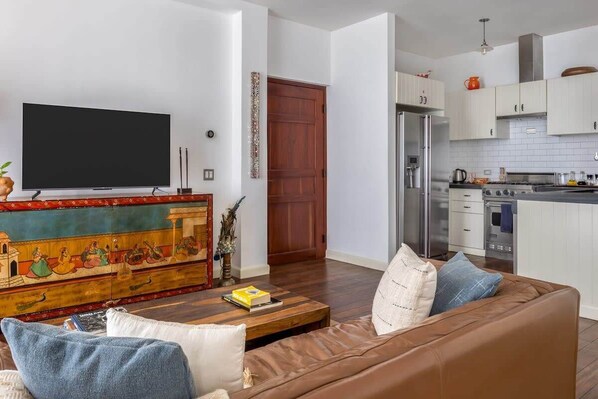 A living room with a brown sofa, a coffee table, and a TV. There is a kitchen with white cabinets and a refrigerator.