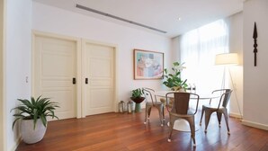 A room with wooden flooring, a dining table with chairs, a potted plant, a wall-mounted lamp, and a framed picture.
