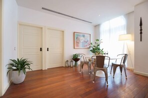 A room with wooden flooring, two white doors, a dining table with four chairs, a potted plant, a wall-mounted lamp, and a framed picture.