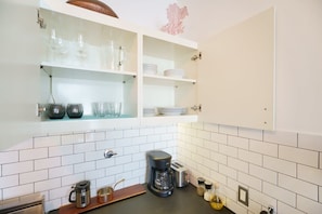 Una cocina con salpicadero de azulejos blancos, cafetera, tostadora y estantes con cristalería y platos.