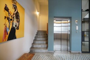 Un pasillo con una escalera que conduce a un ascensor, suelo de baldosas y una pared decorada con un cuadro abstracto de gran tamaño.
