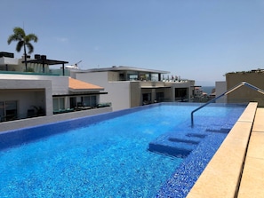 En swimmingpool med en klar, blå vandoverflade, omgivet af moderne bygninger med altaner og en klar himmel.
