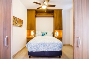 Un dormitorio con cama, ventilador de techo, dos lámparas de noche y un cuadro en la pared.