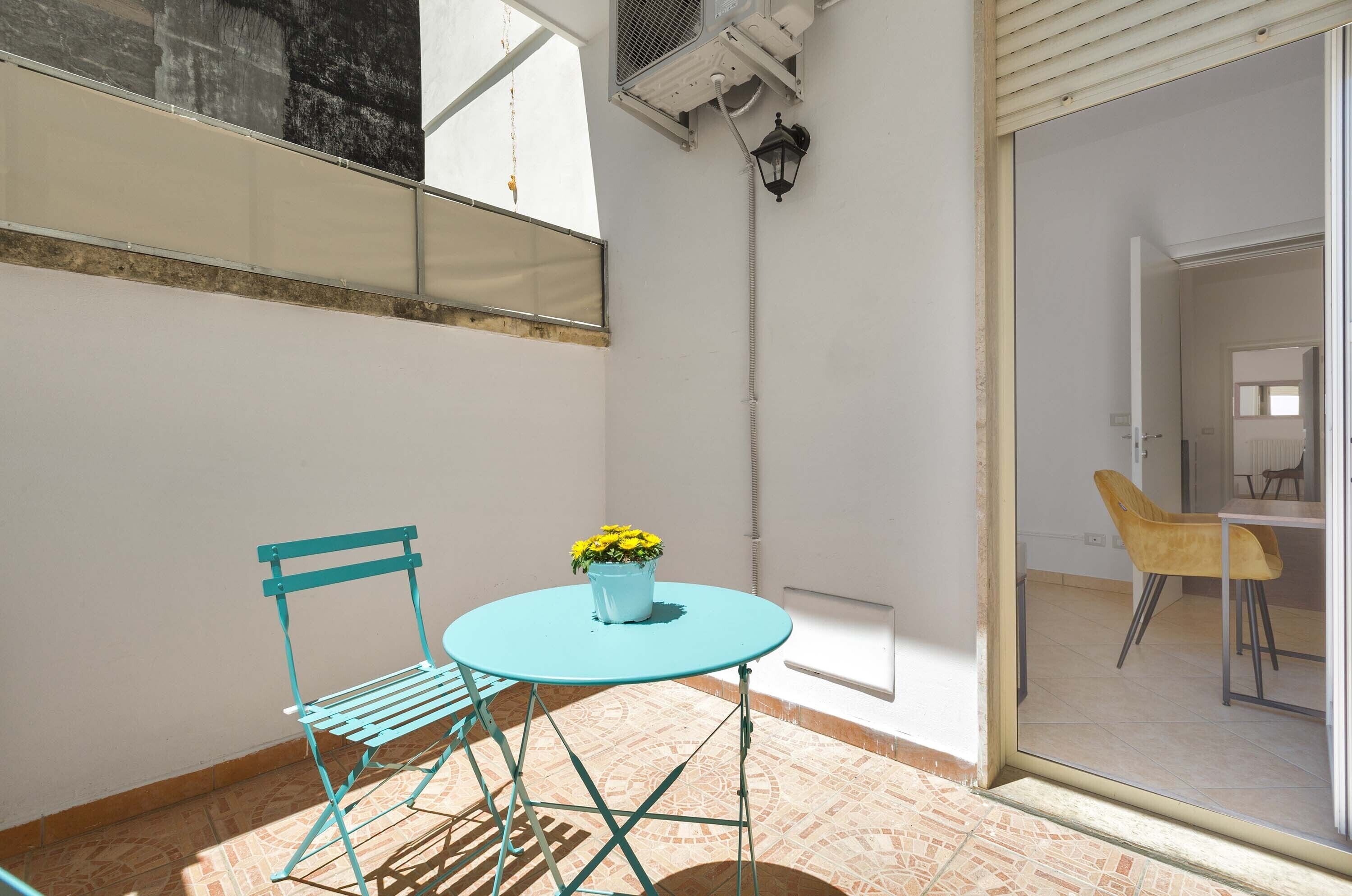 A small balcony with a turquoise metal table and chair, a potted plant, and a view of an adjacent room.