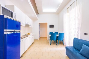 Une cuisine moderne avec des meubles blancs et un réfrigérateur bleu, un coin repas avec des chaises bleues et un canapé bleu dans une pièce lumineuse dotée d’une verrière.