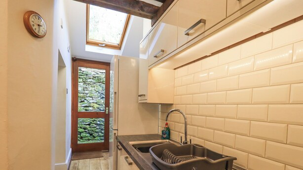 A compact kitchen with a built-in sink, a window above the door, and a view of a stone wall through the glass door. - Slaters Rest (Ambleside)