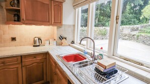 A kitchen with wooden cabinets, a sink with a dish rack, and a window with a view of greenery. - Miyanli (Windermere)