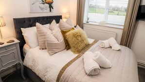 A neatly made bed with a headboard, pillows, and a fluffy throw. There is a painting on the wall above the bed and a window with curtains.