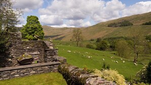 A stone structure with a stone bench and a statue, surrounded by greenery and sheep grazing in the background. - Thyme Out (Windermere)