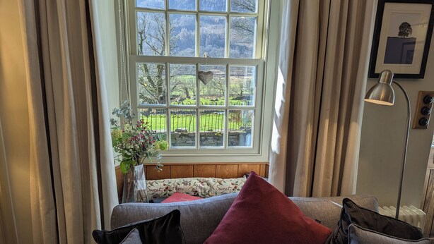 Ein Zimmer mit großem Fenster, einer Couch mit Kissen, einer Vase mit Blumen und einer Lampe.