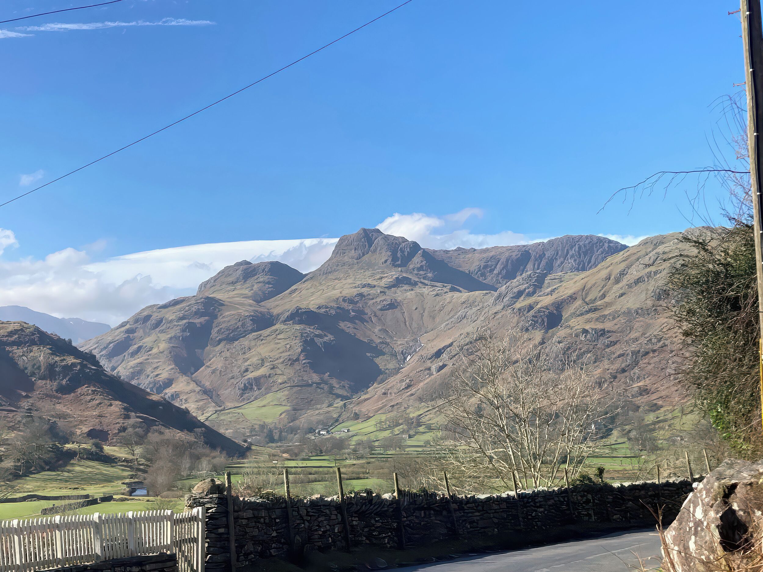 A rural road with stone walls and a wooden fence, leading to a valley with mountains and a clear sky.