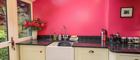 A kitchen with a red wall, a window with a view of greenery, and a sink with various items on the counter.