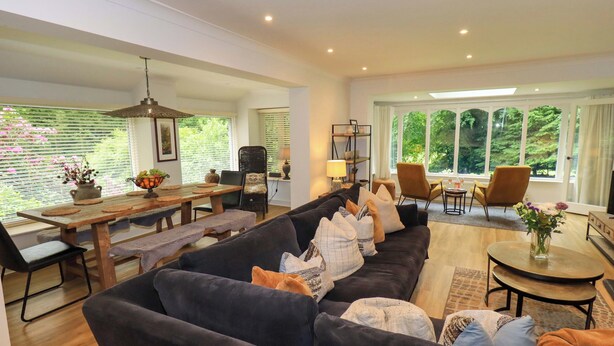 A spacious living area with a dark-colored sofa, wooden dining table, and chairs. There are large windows with a view of greenery outside. - Underheron (Ambleside)