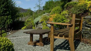 Un banc en bois dans un jardin, avec un chemin de gravier, entouré de différentes plantes et d’un mur de pierre.