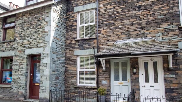 A two-story stone building with a stone facade, white-framed windows, and a black metal fence. - Acorn Cottage (Ambleside)