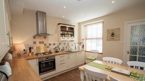 A compact kitchen with a gas stove, oven, and a built-in range hood. There is a dining area with a table and chairs, and a window with blinds. - Acorn Cottage (Ambleside)