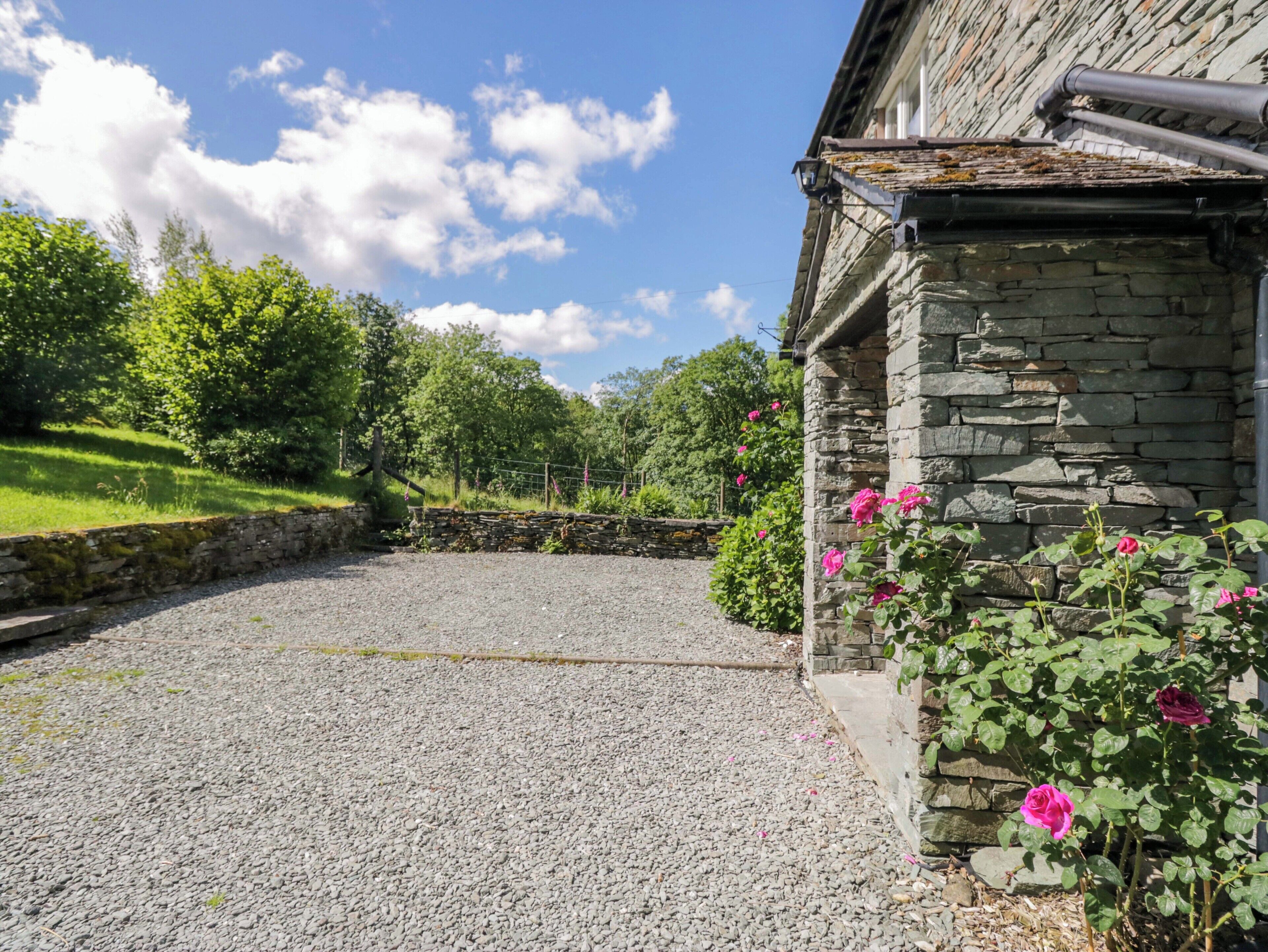 Um edifício de pedra com entrada por um caminho de cascalho, rodeado por vegetação e flores cor-de-rosa.