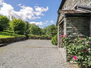 Ein Kiesweg führt zu einem Steinhaus mit einer Steinmauer und einem Blumenbeet mit rosa Blüten.