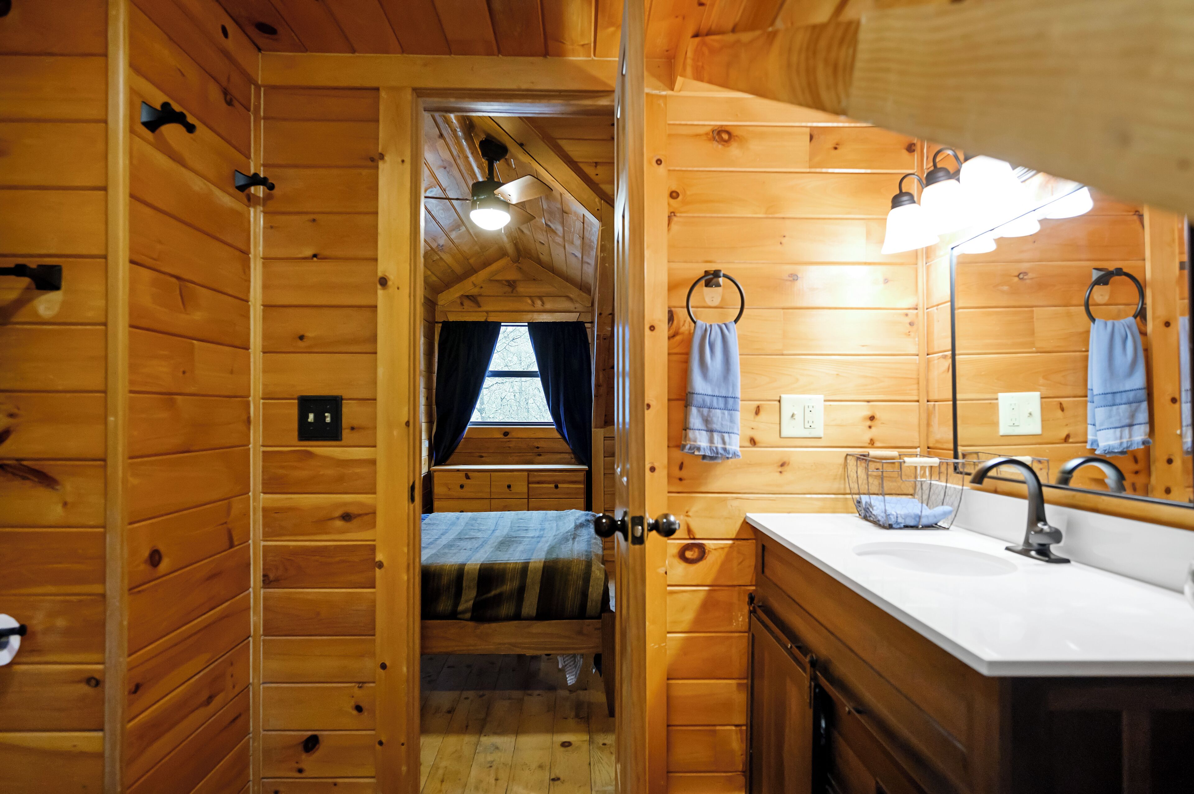 Interior de uma cabana de madeira com um quarto e um banheiro. O quarto possui uma cama com cobertor listrado e uma janela com cortinas. O banheiro tem uma pia, espelho e porta-toalhas.