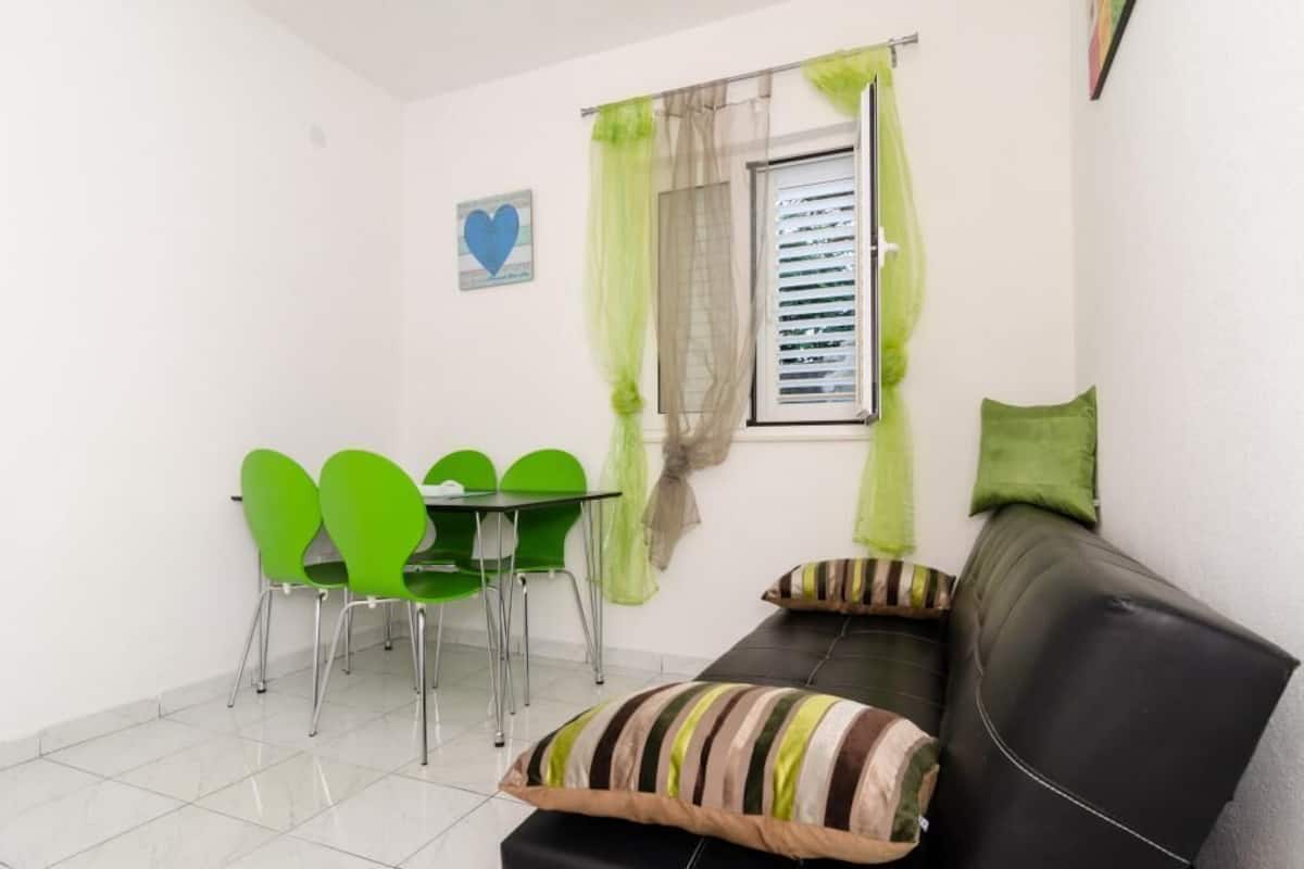 Une petite pièce propre, avec un canapé noir, des chaises vertes, une table et une fenêtre ornée de rideaux légers.