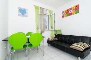 Une pièce avec un canapé noir, des fauteuils verts, une petite table, une fenêtre avec des rideaux et un élément de décoration murale aux couleurs vives.