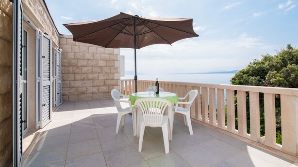 Ein Balkon mit Tisch und Stühlen, einem großen Sonnenschirm und Blick aufs Meer.
