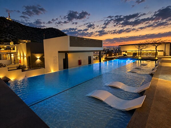 A swimming pool with lounge chairs, a building with a flat roof, and a sunset view.