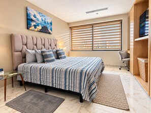 A bedroom with a bed, a desk, a chair, a TV, and a window with blinds.