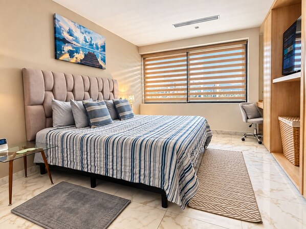 A bedroom with a bed, a desk, a chair, a TV, and a window with blinds.