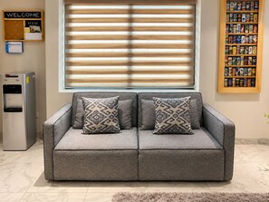 A grey sofa with patterned cushions, a water dispenser, a welcome sign, and a beverage shelf.